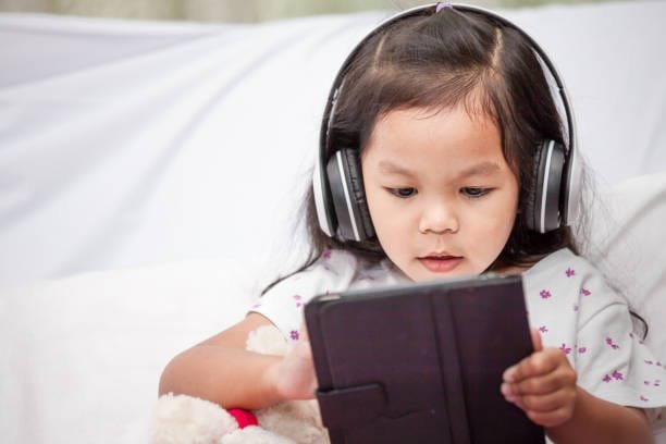 cute asian little child girl in headphones is using a tablet in her room